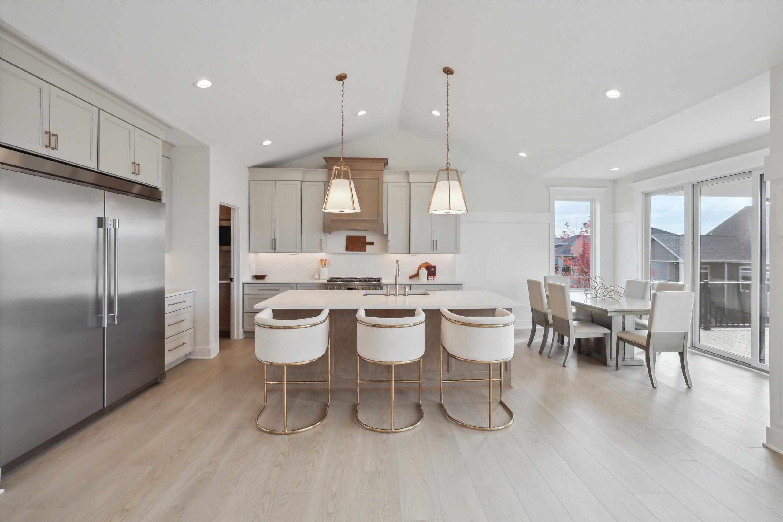 A modern kitchen features a large island with four white and gold stools, overhead pendant lights, and built-in stainless steel appliances. Adjacent is a dining area with a table and chairs by large windows letting in natural light.