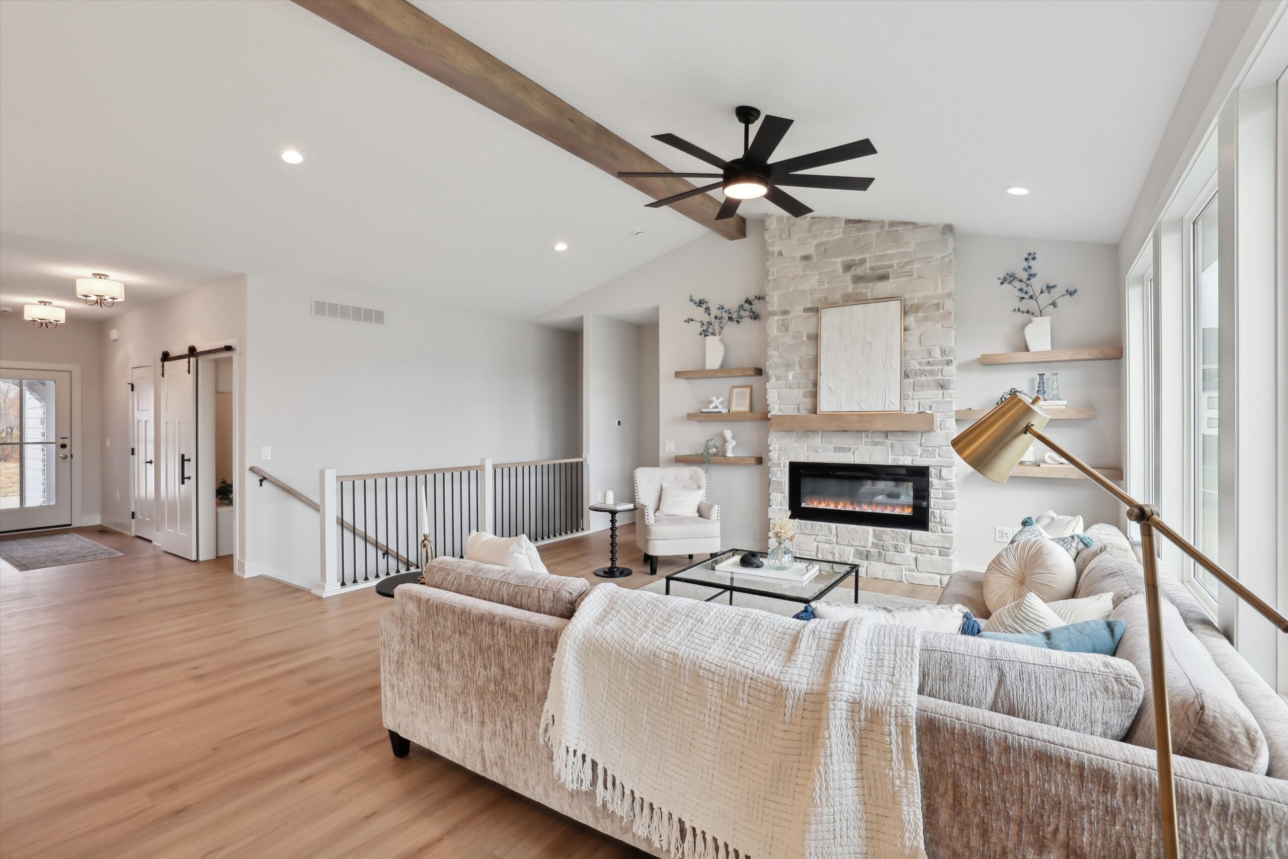 A modern living room with a beige sofa, white throw, and cushions facing a stone fireplace. The room features light wood flooring, large windows, and a ceiling fan. A floor lamp stands nearby, and a sliding barn door is visible in the hallway.