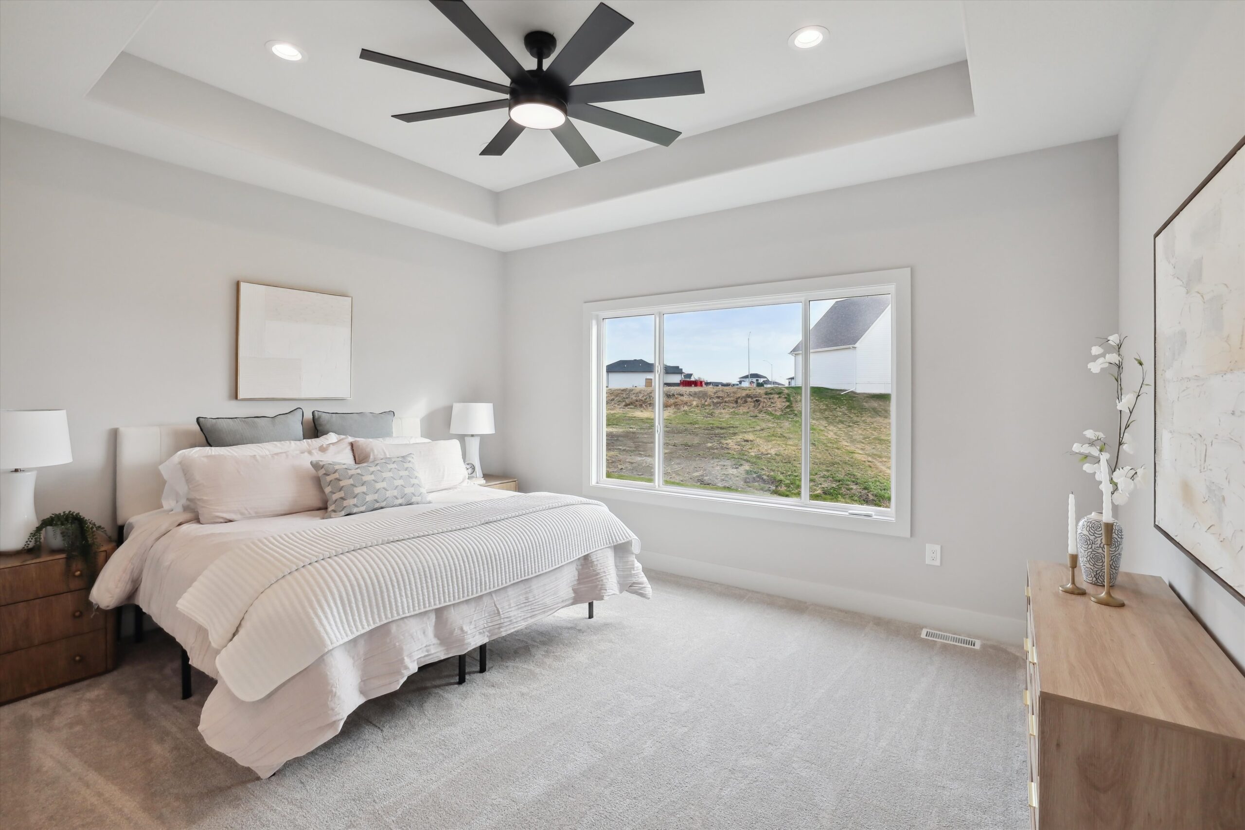 A modern bedroom featuring a large bed with white and gray bedding, flanked by two nightstands and lamps. A ceiling fan hangs above, and a large window offers a view of the outdoors. Neutral tones dominate the decor.