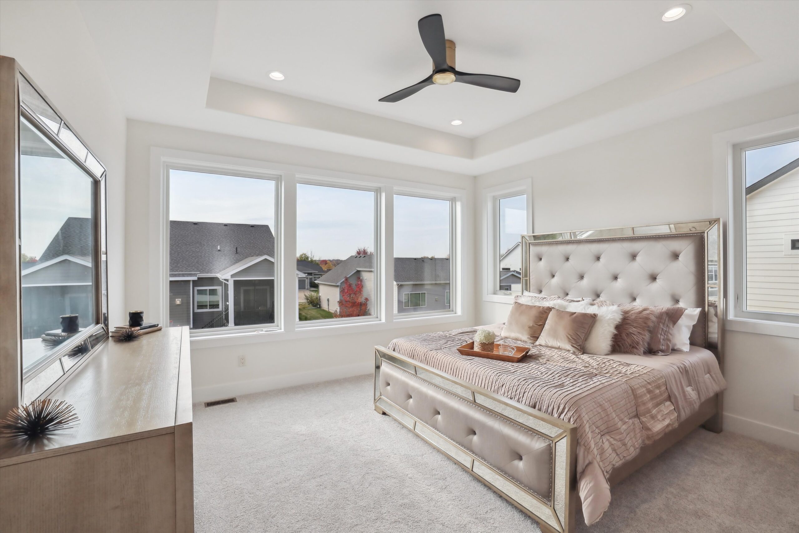 A bright bedroom with a large bed featuring a tufted headboard, mirrored frame, and beige bedding. A tray is placed on the bed. Large windows surround the room, providing natural light. A ceiling fan hangs above, and a wooden dresser sits opposite the bed.