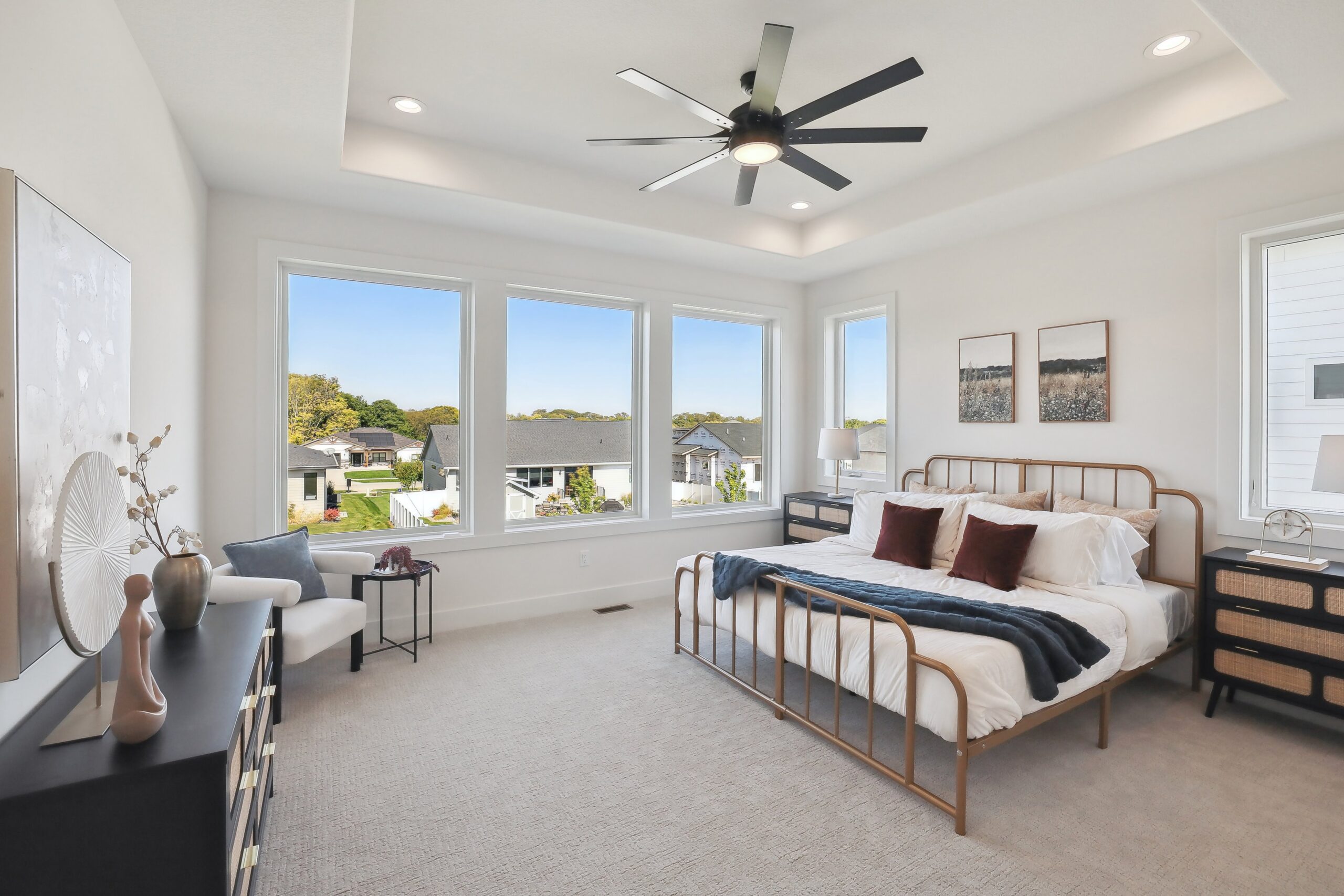 A bright, modern bedroom with a large bed, decorative pillows, a dresser, and armchair. Large windows let in natural light and offer a view of houses and greenery outside. A ceiling fan is centered on a recessed ceiling.