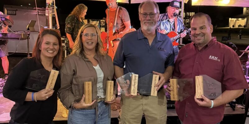 Four people stand under a tent, smiling and holding plaques. A man with a guitar and other band members perform on stage behind them.