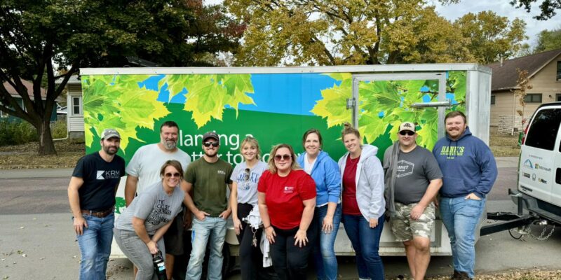 A group of people standing in front of a trailer with green and blue graphics, depicting leaves and trees. They are outdoors on a grassy area, with trees in the background. Everyone is dressed casually, smiling, and posing for the photo.