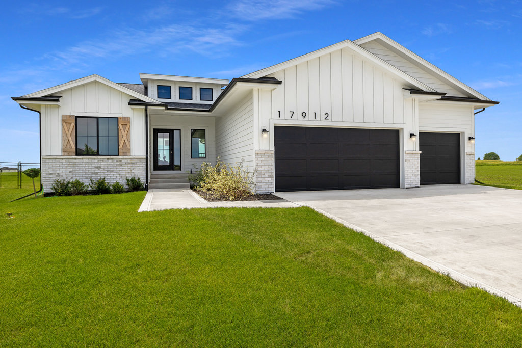 A modern, white farmhouse-style home with a black front door and three-car garage set against a clear blue sky. The house has a large lawn and a paved driveway leading to the entrance.