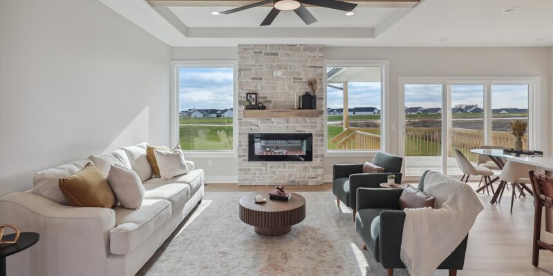 Modern living room with white sofa, two green armchairs, and a round coffee table. A stone fireplace under a black ceiling fan is centered against a wall with large windows, providing a view of a neighborhood and a lawn. Dining area is visible in the back.