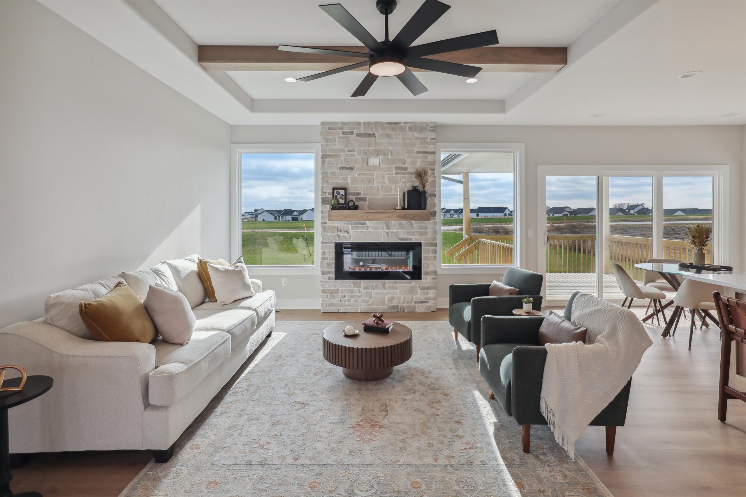 A modern living room with a large ceiling fan, stone fireplace, and large windows overlooking a suburban neighborhood. It features a sofa, two armchairs, a round coffee table, and a dining area in the background. Neutral tones create a cozy atmosphere.