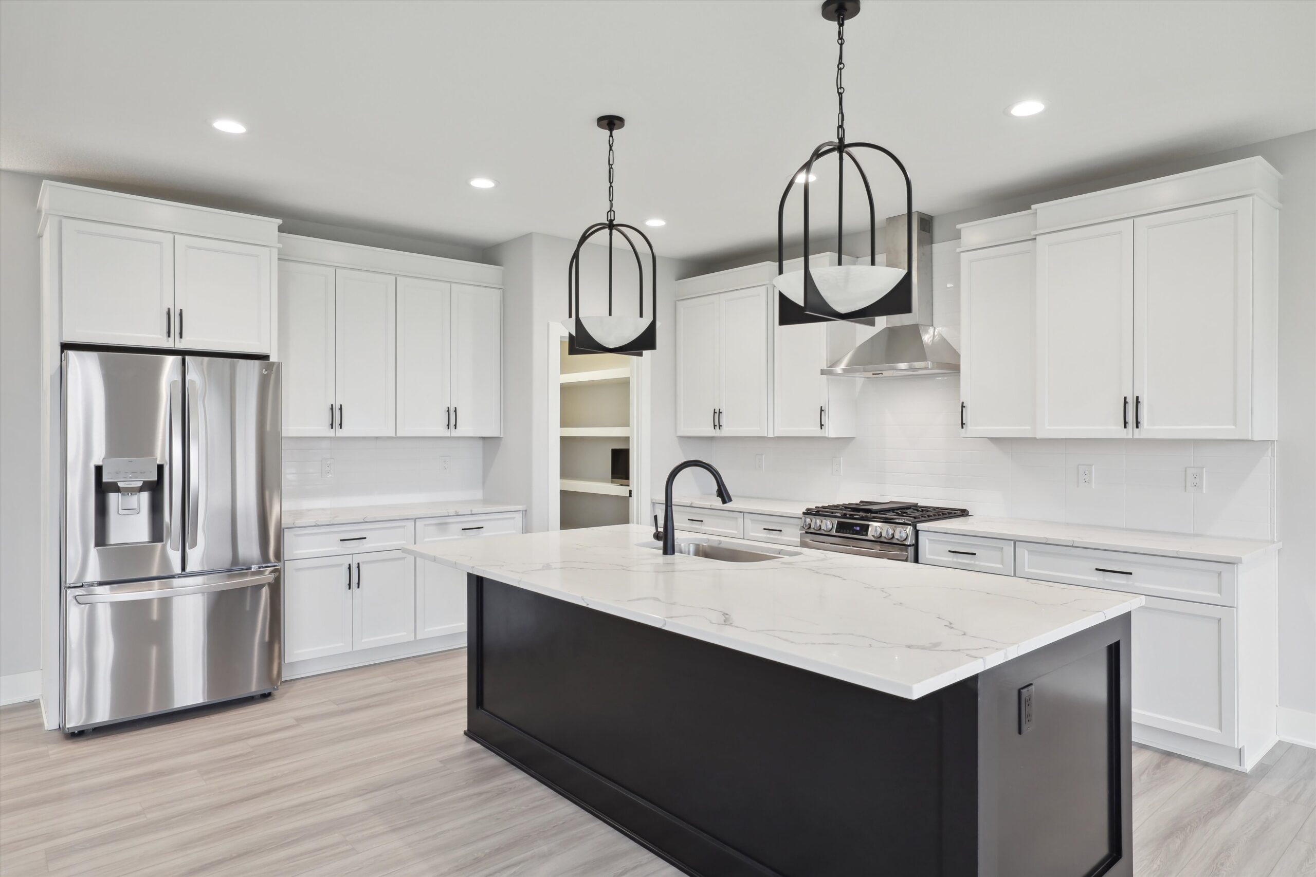 A modern kitchen with white cabinets, a stainless steel refrigerator, and a large marble island. Two black pendant lights hang over the island, which features a black base and a built-in sink. Light wood flooring complements the bright, open space.