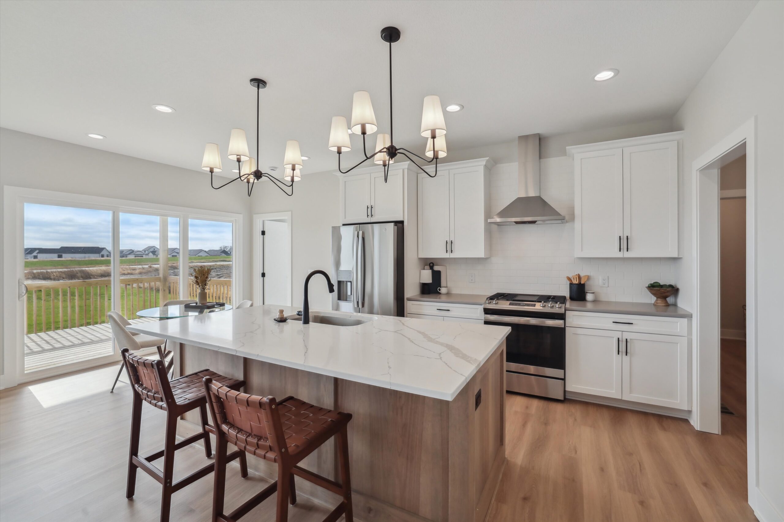 A modern kitchen with white cabinets, a marble island, and brown woven bar stools. A black sink and faucet sit on the island, illuminated by a chandelier. Stainless steel appliances and a view of a deck and field are visible through large windows.