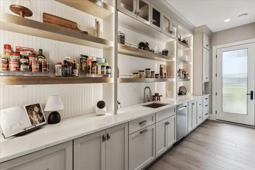 A kitchen wall features long open shelves that display decor and jars of food