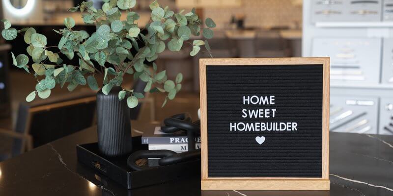 A letter board reads Home Sweet Homebuilder next to a vase with green leaves on a black marble table.
