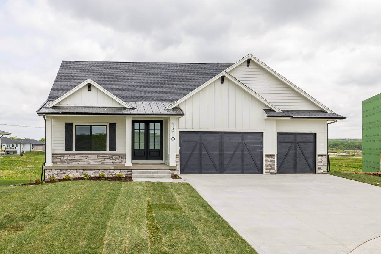 A modern single-story house with a dark gray roof, black trim windows, white siding, and a large double garage with dark doors. The house has a neatly manicured lawn and a concrete driveway. The sky is overcast in the background.