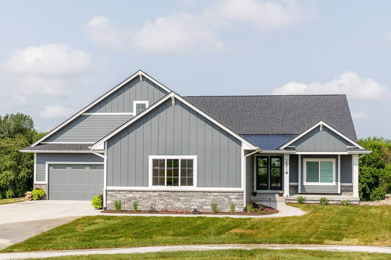 A modern, single-story house with gray siding, a dark gray shingle roof, and white trim. It features a two-car garage, a small front porch, and a well-maintained lawn with young landscaping. The sky is partly cloudy.