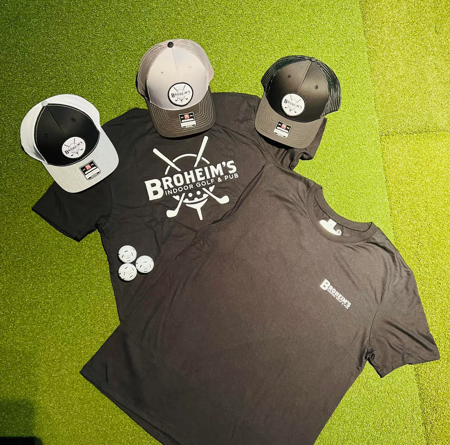 Three hats with logo, two t shirts with logo, and three golf balls placed on a grass surface.