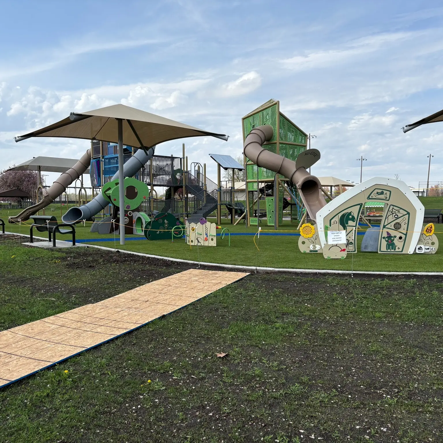 Outdoor playground area. Large play structures with platforms and railings. Multiple slides, including a long enclosed tube slide and an open slide. Shade canopy over part of the structure. Climbing elements and panels attached to the play area. Small activity panels in the front. Artificial turf ground. Pathway leading toward the playground. Benches on the side. Open sky and grassy area around.