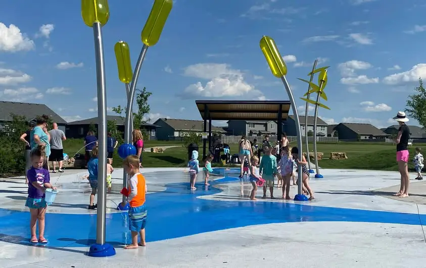 Outdoor play area with water features where children are running and playing around spray structures and shallow ground fountains. The space includes an open layout with nearby seating and a covered area in the background for shade and gathering.