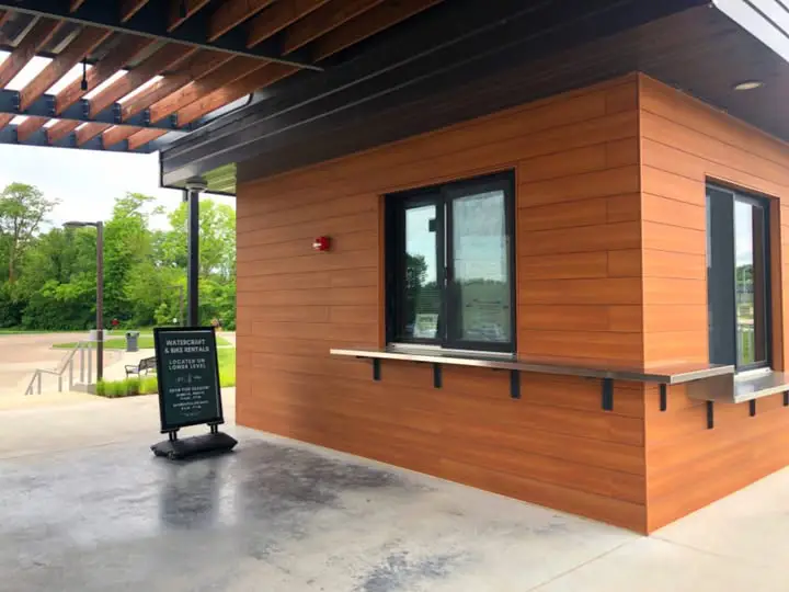 Small outdoor service window attached to a building with a counter underneath. There is a sign nearby and an open area around it for people to walk or wait.
