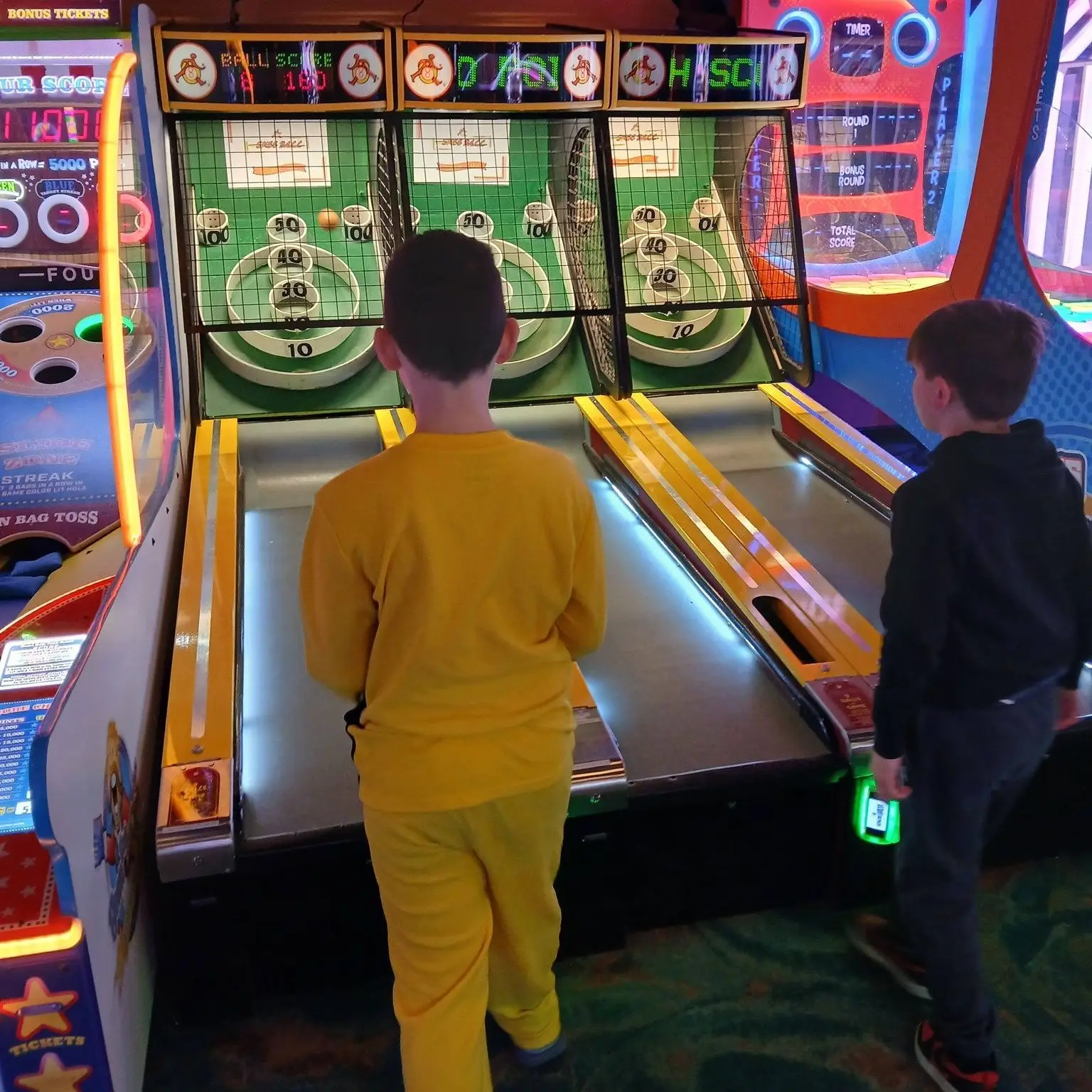 Two kids playing a skee-ball style arcade game, aiming balls up the lanes toward circular scoring targets. Bright arcade lights and score displays are visible above the machines.