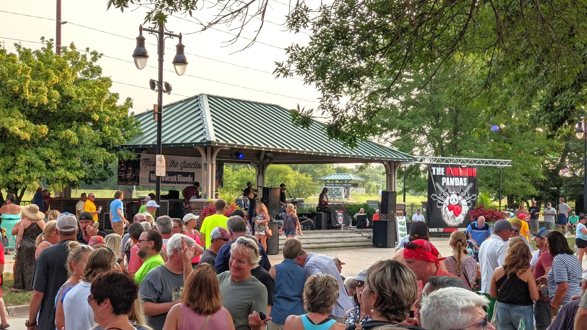 Outdoor live music event with a crowd gathered around a covered stage, featuring a band performing and a lively, festival style atmosphere.
