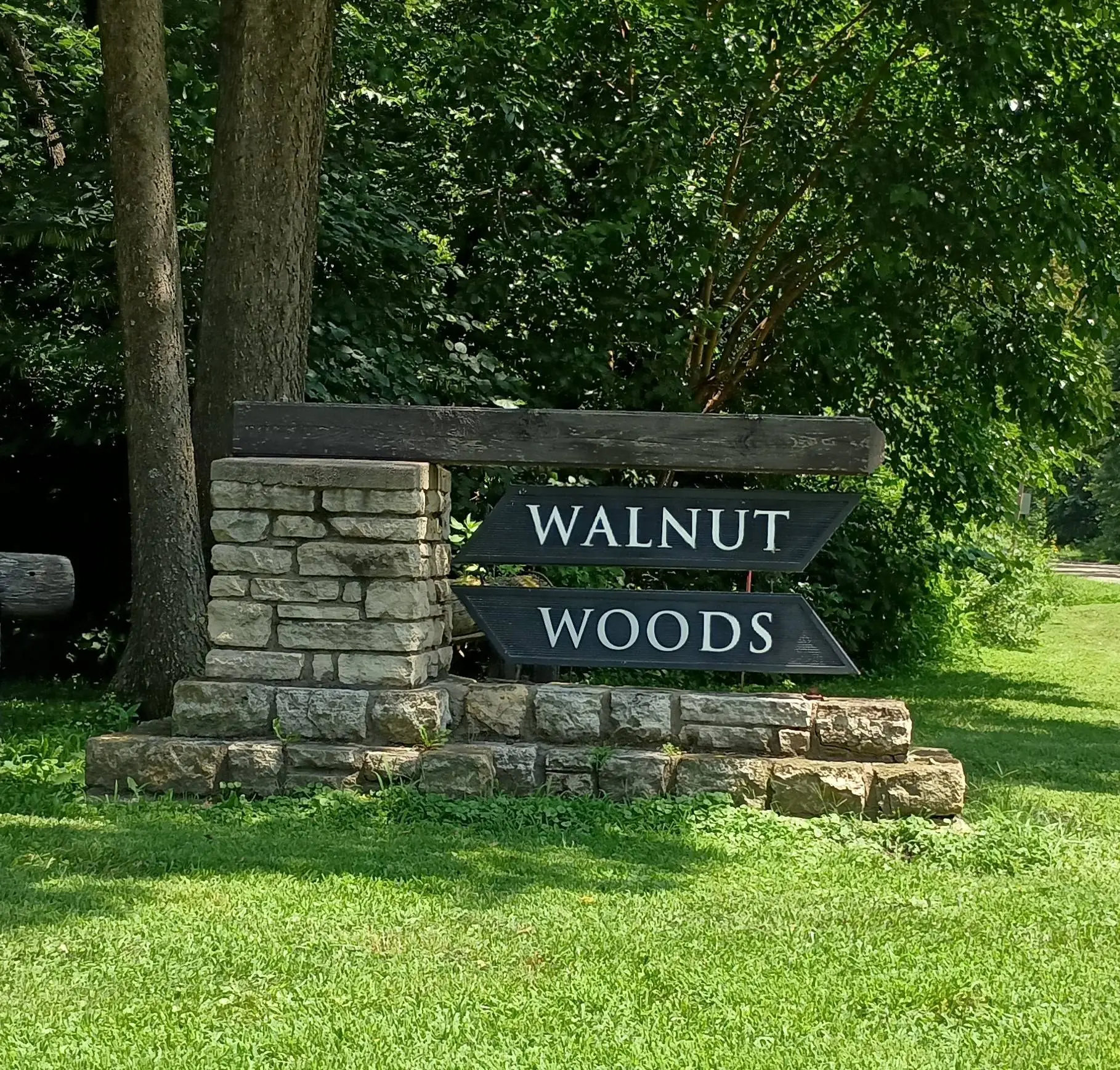 Stone and wood entrance sign reading “Walnut Woods,” set beside a grassy area and surrounded by trees, marking the entrance to a residential neighborhood.