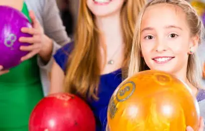 Two girls holding bowling balls. One is holding an orange ball in front. Another is holding a red ball. A third person is partially visible in the background holding a purple ball.