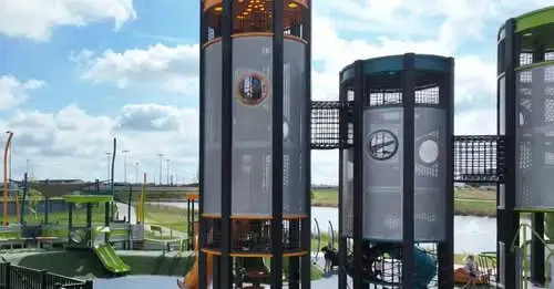 Playground. Tall play structures with slides. Climbing sections and platforms. Outdoor setting with grass and sky.