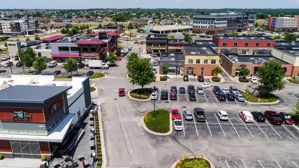 Aerial view of a busy commercial area with multiple retail and restaurant buildings, large parking lots, and steady car traffic. The layout shows a well-developed neighborhood hub with shops, dining spots, and nearby residential areas, making it convenient for everyday errands and social activity.