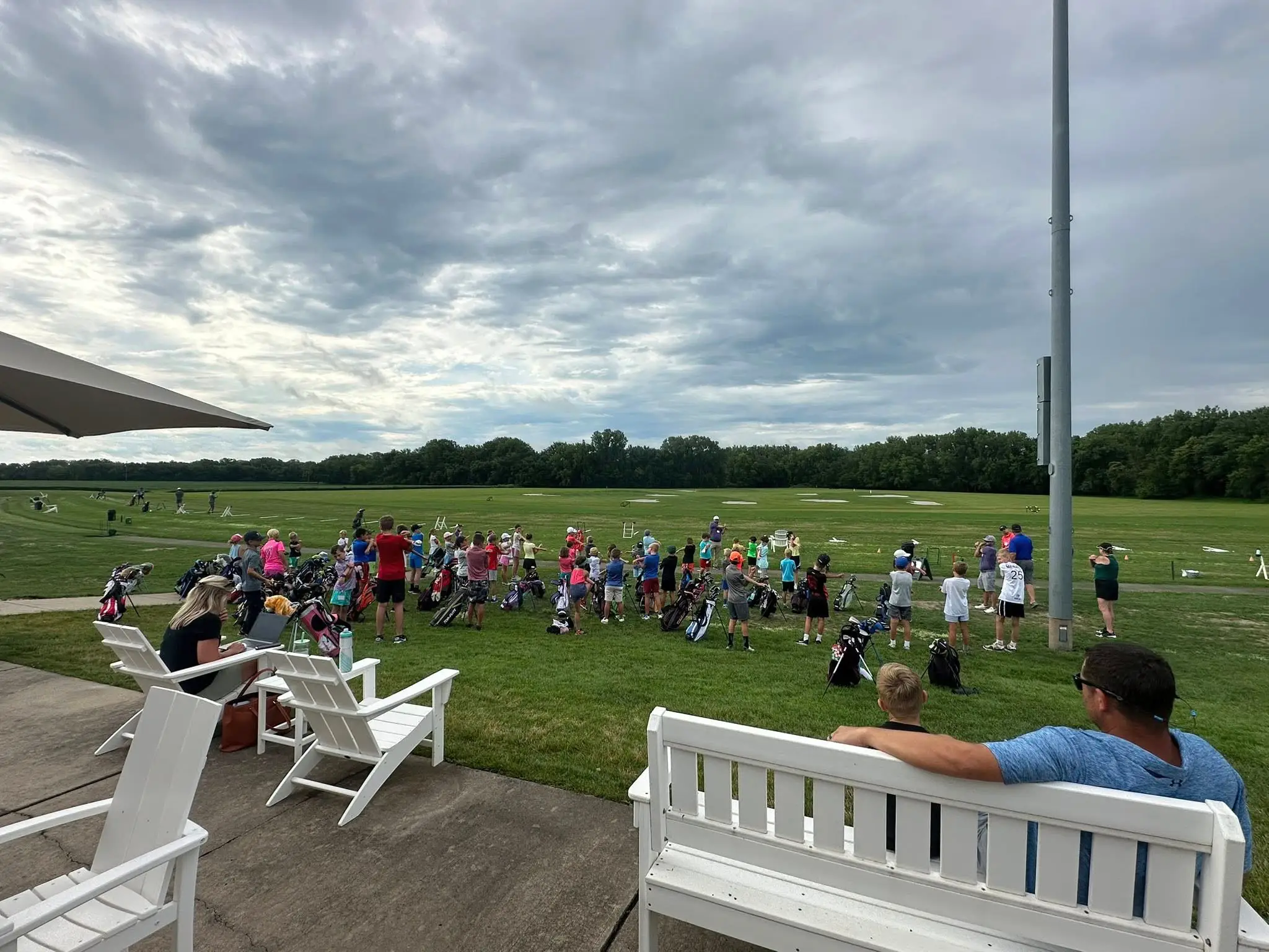 Outdoor golf practice area with a large group of people lined up on the range. Many are holding clubs or standing near golf bags, while others watch from seating behind. The open field stretches out with targets in the distance, and the scene feels like a group lesson or event.